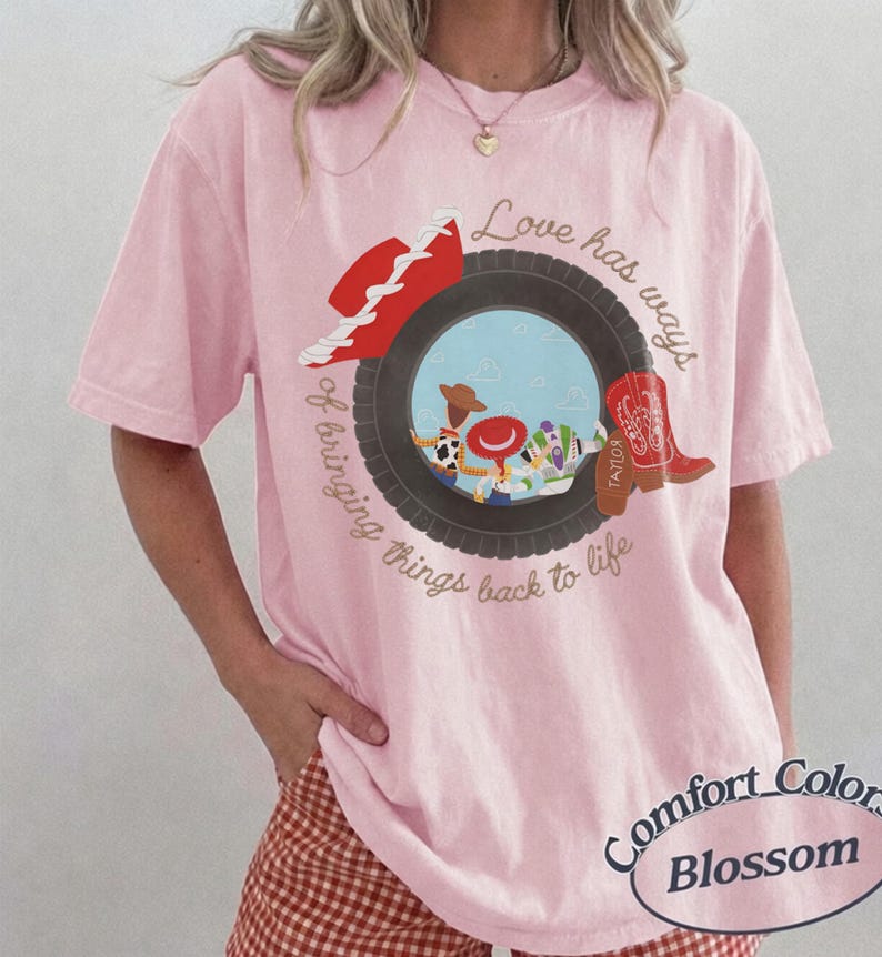 May include: A light pink short-sleeve t-shirt featuring a graphic of a black tire framing cartoon characters. The design includes a red hat, a red cowboy boot with the name Taylor, and the text Love has ways of bringing things back to life.
