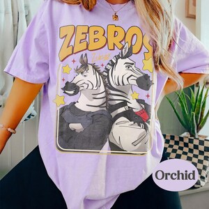 Op de afbeelding: Lichtpaars T-shirt met het woord "ZEBROS" in geel en een cartoonafbeelding van twee zebra's. De zebra's dragen kleding en zijn omringd door sterren. Het woord "Orchid" staat rechtsonder.