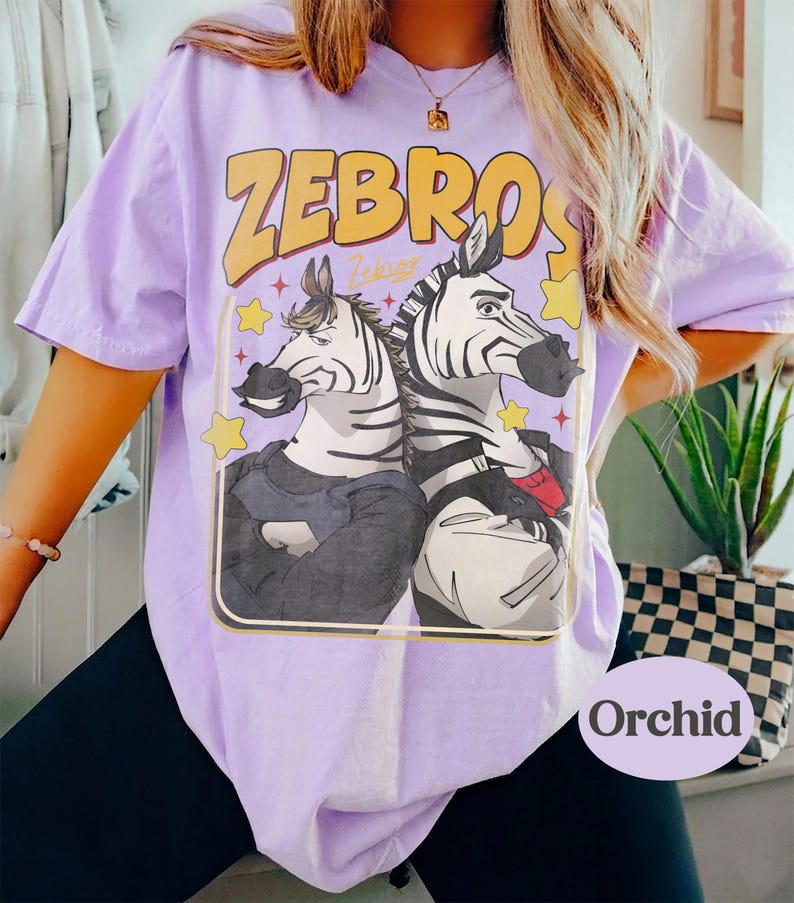 Op de afbeelding: Lichtpaars T-shirt met het woord "ZEBROS" in geel en een cartoonafbeelding van twee zebra's. De zebra's dragen kleding en zijn omringd door sterren. Het woord "Orchid" staat rechtsonder.