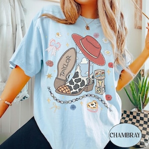 Puede incluir: Una camiseta azul claro con un estampado gr&aacute;fico de una bota de vaquero con estampado de vaca con el nombre Taylor, un sombrero de vaquero rojo, bloques de letras que deletrean TS, un cord&oacute;n de cuentas y varios iconos florales y de estrellas.