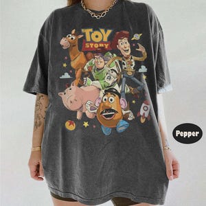 May include: A charcoal gray short-sleeve t-shirt featuring a vintage-style graphic print of Toy Story characters including Woody, Buzz Lightyear, Bullseye, Hamm, and Mr. Potato Head, with the Toy Story logo and colorful stars and planets.