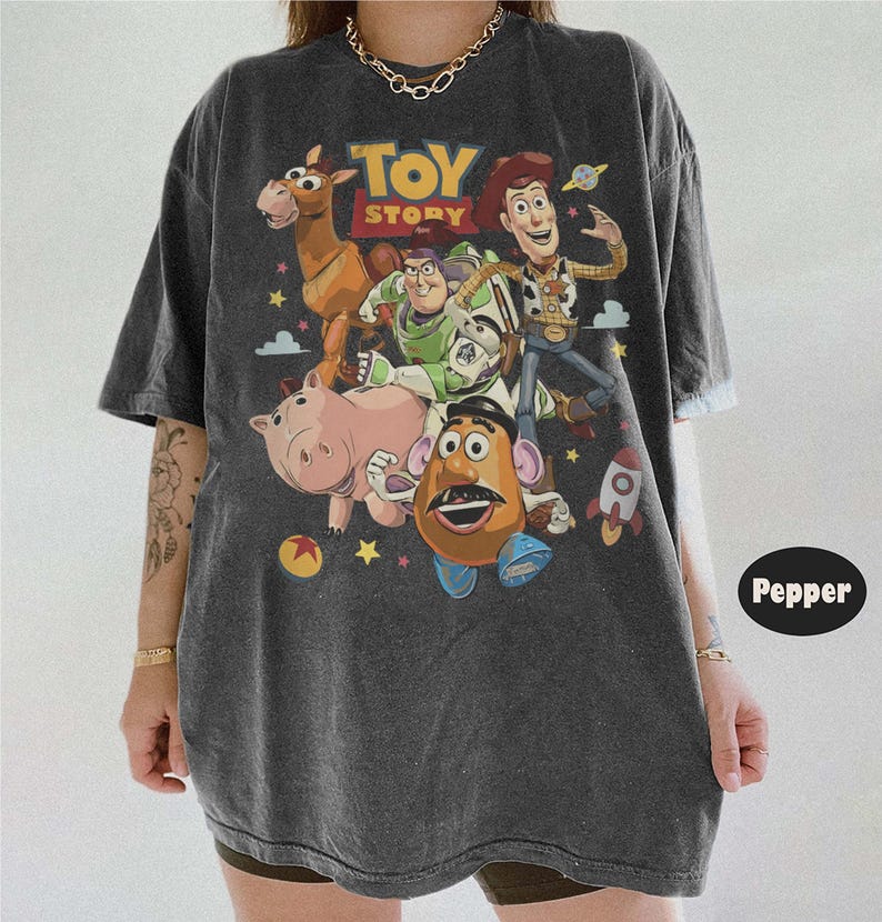May include: A charcoal gray short-sleeve t-shirt featuring a vintage-style graphic print of Toy Story characters including Woody, Buzz Lightyear, Bullseye, Hamm, and Mr. Potato Head, with the Toy Story logo and colorful stars and planets.