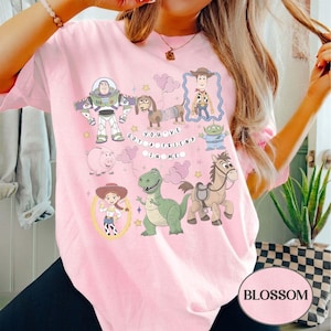 Pu&ograve; includere: T-shirt rosa chiaro con personaggi dei cartoni animati del film Toy Story. Il design include Buzz Lightyear, Woody, Jessie, Rex e altri personaggi, con la scritta "You've Got a Friend in Me."