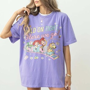 May include: A lilac-colored t-shirt featuring a graphic of Toy Story characters on a roller coaster. The text "HOLD ON TIGHT, Here we go!" is printed above the image. The shirt has short sleeves and a crew neck.