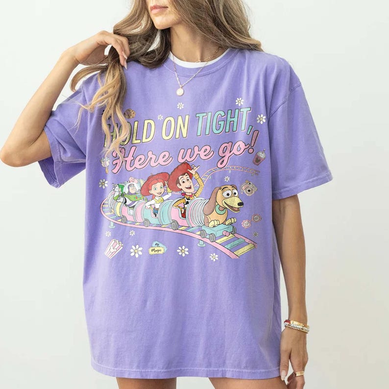 May include: A lilac-colored t-shirt featuring a graphic of Toy Story characters on a roller coaster. The text "HOLD ON TIGHT, Here we go!" is printed above the image. The shirt has short sleeves and a crew neck.
