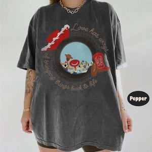 May include: A charcoal gray short-sleeve t-shirt featuring a graphic of a tire framing characters from Toy Story. The text reads Love has ways of bringing things back to life. A red cowboy hat and a red cowboy boot are positioned on the tire.