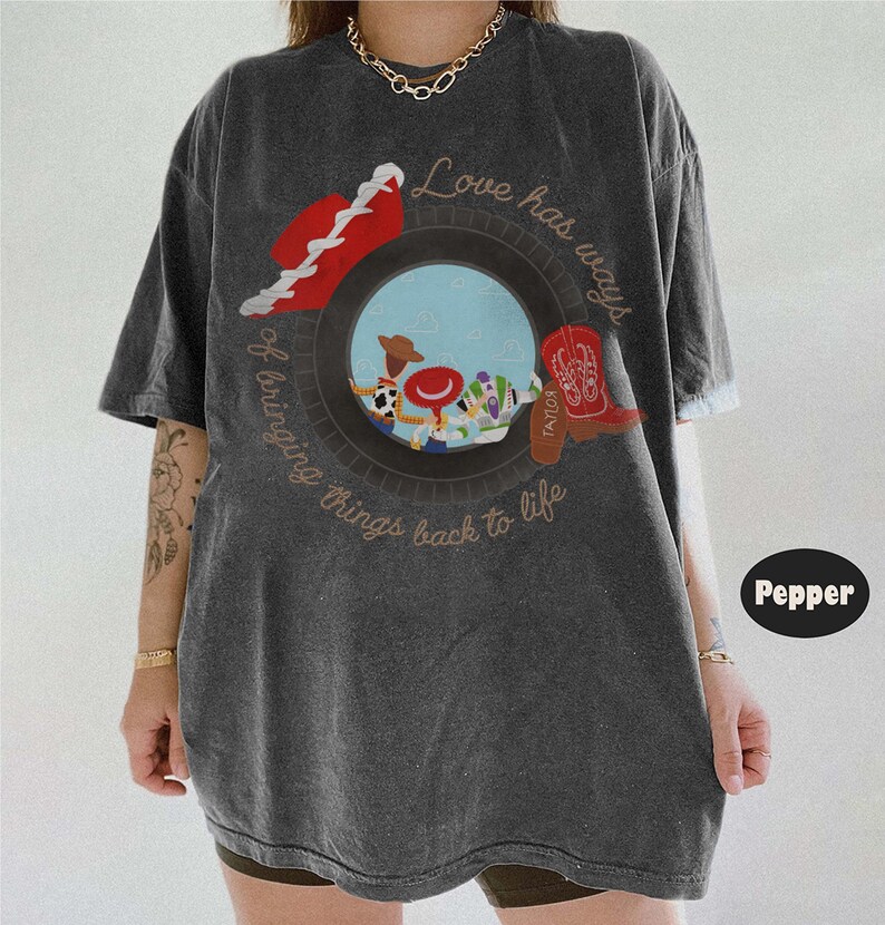 May include: A charcoal gray short-sleeve t-shirt featuring a graphic of a tire framing characters from Toy Story. The text reads Love has ways of bringing things back to life. A red cowboy hat and a red cowboy boot are positioned on the tire.