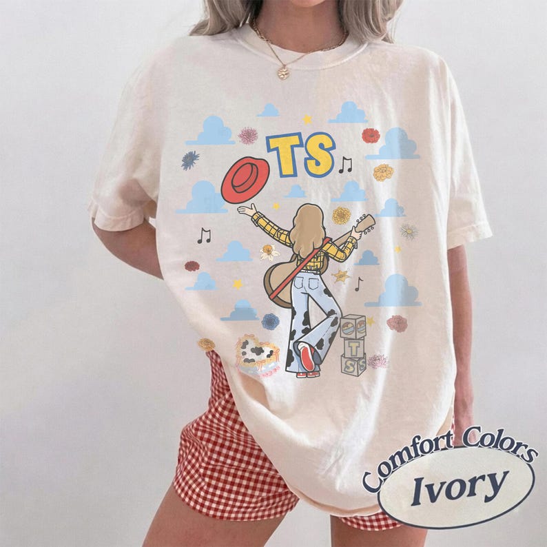 Op de afbeelding: Een ivoorkleurig T-shirt met korte mouwen en een afbeelding van een persoon met lang haar die gitaar speelt, gekleed in een geel geruit overhemd en een broek met koeienprint. Het ontwerp bevat blauwe wolken, muzieknoten en de tekst TS.