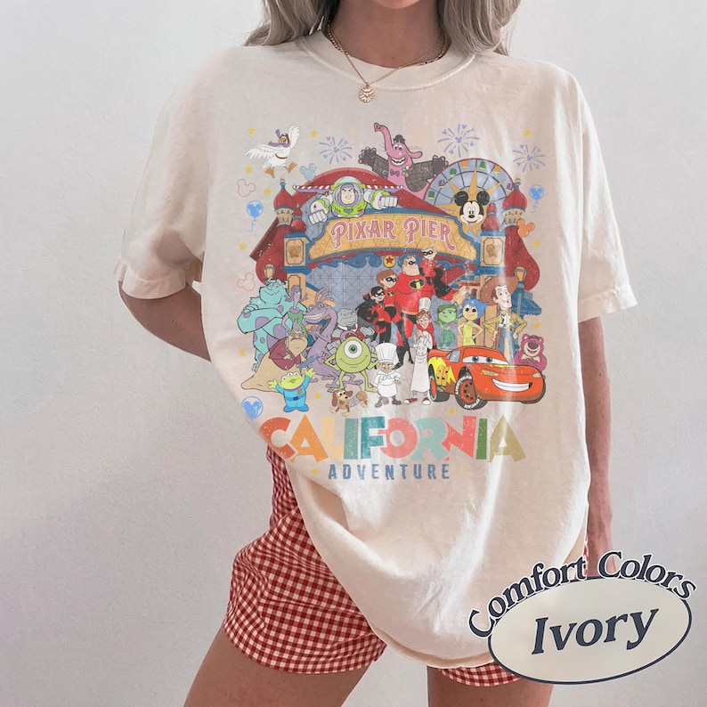 May include: Ivory-colored t-shirt featuring a colorful graphic of Pixar Pier characters and the words "California Adventure." The shirt is a Comfort Colors brand.