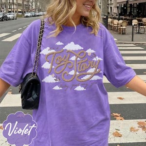 Pu&ograve; includere: Una t-shirt viola a maniche corte con una grafica di nuvole bianche e il testo "it's a Toy Story baby, just say yes" scritto in un carattere color marrone chiaro che ricorda una corda. Una borsa a tracolla nera trapuntata con catena dorata.