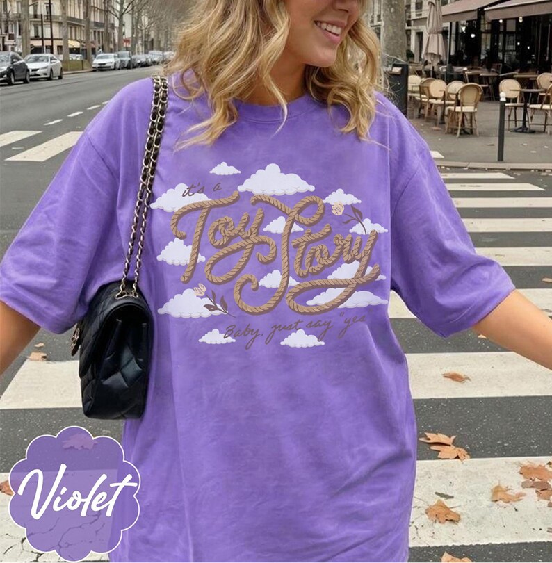 Pu&ograve; includere: Una t-shirt viola a maniche corte con una grafica di nuvole bianche e il testo "it's a Toy Story baby, just say yes" scritto in un carattere color marrone chiaro che ricorda una corda. Una borsa a tracolla nera trapuntata con catena dorata.