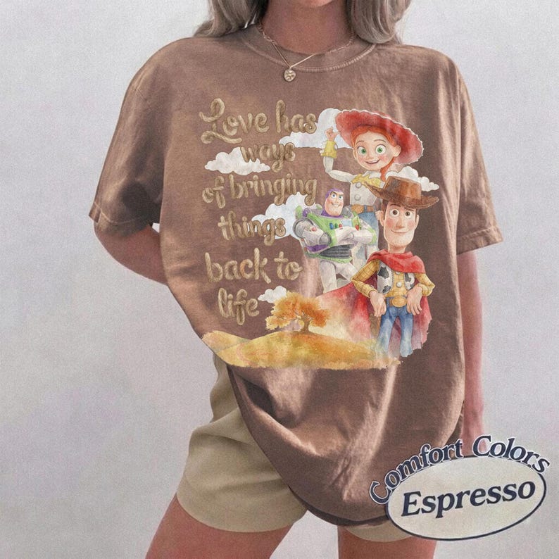 May include: A brown short-sleeved t-shirt featuring a graphic print of Woody, Buzz Lightyear, and Jessie from Toy Story. The text reads, Love has ways of bringing things back to life. The design includes clouds and a small tree on a hill.
