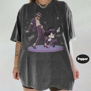 Pode incluir: Uma t-shirt cinzento-carv&atilde;o oversized com uma impress&atilde;o gr&aacute;fica de Michael Jackson e Mickey Mouse, ambos a usar fatos e chap&eacute;us fedora roxos. O design inclui pequenas estrelas brancas e as assinaturas de ambas as figuras num palco circular roxo.