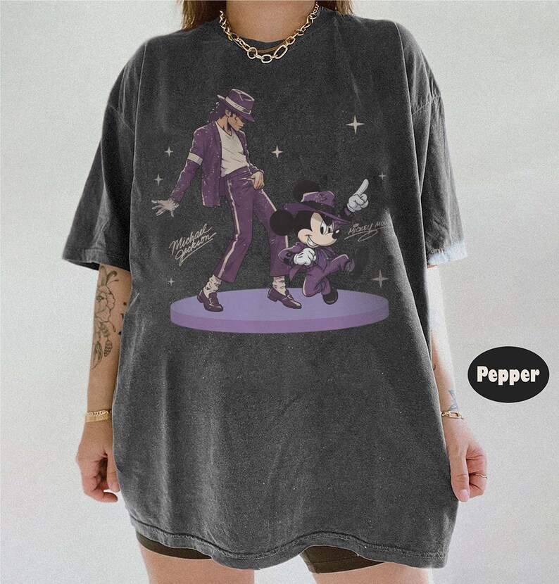 Pode incluir: Uma t-shirt cinzento-carv&atilde;o oversized com uma impress&atilde;o gr&aacute;fica de Michael Jackson e Mickey Mouse, ambos a usar fatos e chap&eacute;us fedora roxos. O design inclui pequenas estrelas brancas e as assinaturas de ambas as figuras num palco circular roxo.