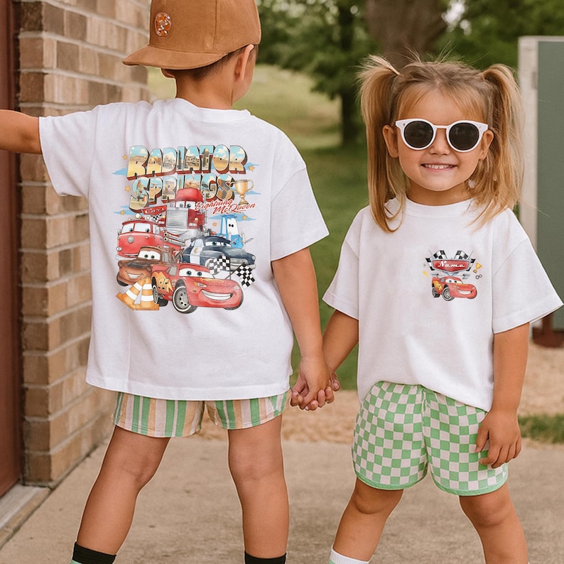 May include: Two children wearing white t-shirts with Cars movie graphics. One shirt features a large design on the back with the text "Radiator Springs" and various car characters. The other shirt has a smaller design on the front pocket area. One child wears green checkered shorts.