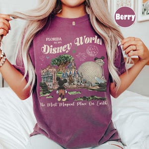 Może przedstawiać: Koszulka w kolorze jagodowym z grafiką Disney World na Florydzie. Projekt zawiera tekst "Florida Disney World Est. 1971" i "The Most Magical Place On Earth". Grafika przedstawia charakterystyczne punkty parku i Myszka Miki.