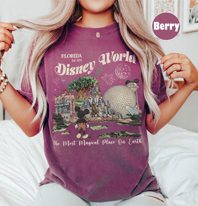 Może przedstawiać: Koszulka w kolorze jagodowym z grafiką Disney World na Florydzie. Projekt zawiera tekst "Florida Disney World Est. 1971" i "The Most Magical Place On Earth". Grafika przedstawia charakterystyczne punkty parku i Myszka Miki.