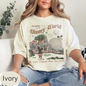 Może przedstawiać: Kremowa koszulka z grafiką Disney World. Projekt zawiera napisy "Florida Disney World" i "The Most Magical Place On Earth", a także ilustracje Myszki Miki, zamku i innych punkt&oacute;w orientacyjnych parku.