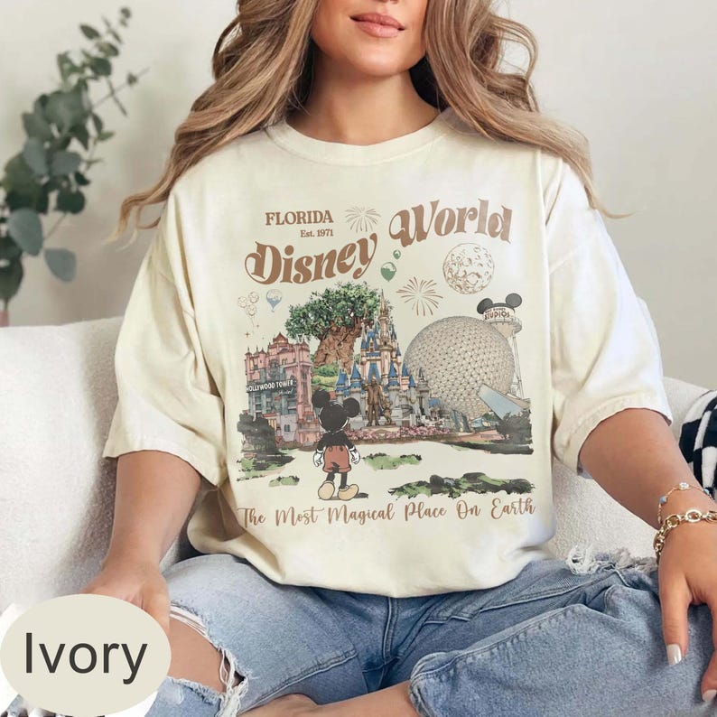 Może przedstawiać: Kremowa koszulka z grafiką Disney World. Projekt zawiera napisy "Florida Disney World" i "The Most Magical Place On Earth", a także ilustracje Myszki Miki, zamku i innych punkt&oacute;w orientacyjnych parku.