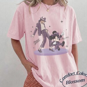 Pode incluir: Uma t-shirt de manga curta rosa claro com uma estampa gr&aacute;fica de Michael Jackson e Mickey Mouse a dan&ccedil;ar numa plataforma circular roxa. O design inclui pequenos detalhes de estrelas e o texto Michael Jackson e Mickey Mouse.