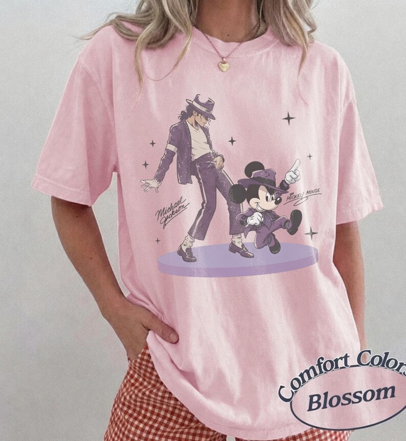 Pode incluir: Uma t-shirt de manga curta rosa claro com uma estampa gr&aacute;fica de Michael Jackson e Mickey Mouse a dan&ccedil;ar numa plataforma circular roxa. O design inclui pequenos detalhes de estrelas e o texto Michael Jackson e Mickey Mouse.