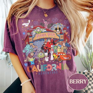 May include: Berry-colored t-shirt featuring a colorful graphic of Pixar Pier characters and the words "CALIFORNIA ADVENTURE." The design includes characters from various Pixar films, such as Buzz Lightyear, Mickey Mouse, and Lightning McQueen.