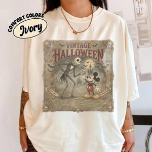 May include: An ivory t-shirt featuring a graphic print with the text 'VINTAGE HALLOWEEN' and 'EST. 1937 CELEBRATION'. The illustration depicts a tall skeletal figure and a cartoon mouse standing in a graveyard with bats, a castle, and skulls.