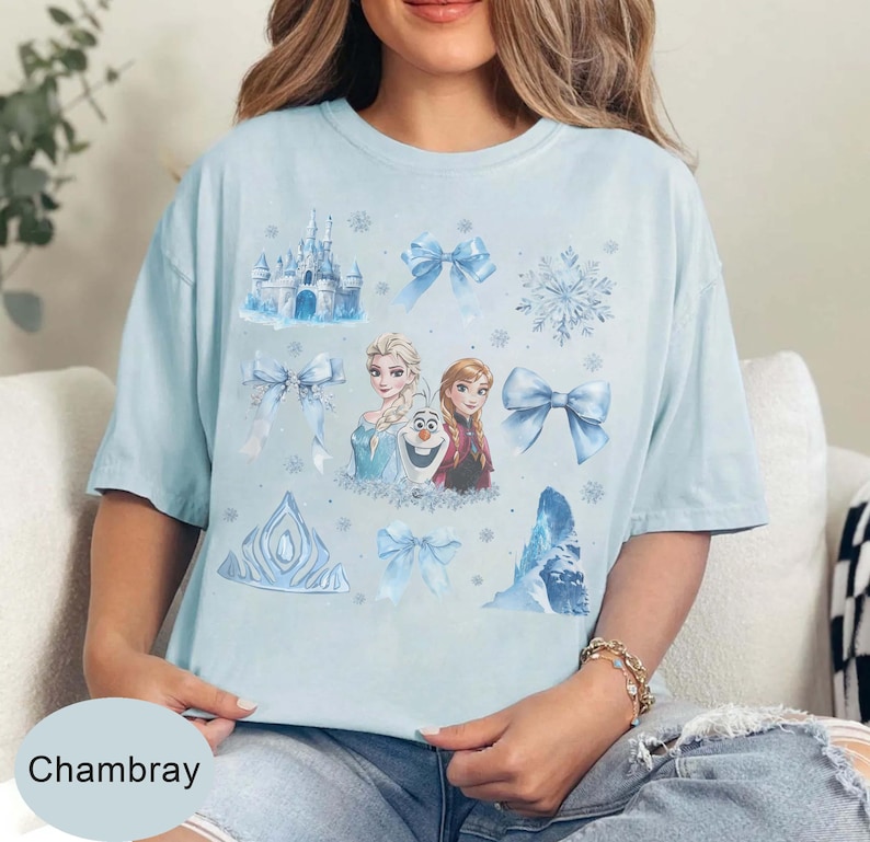 May include: Pale blue t-shirt with a design featuring characters and elements from a popular animated film. The design includes a castle, bows, snowflakes, and the text "Chambray".
