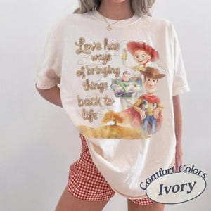 May include: An ivory t-shirt featuring a graphic print of Woody, Buzz Lightyear, and Jessie from Toy Story. The text reads, Love has ways of bringing things back to life, accompanied by a watercolor-style illustration of a tree on a hill.