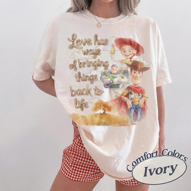 May include: An ivory t-shirt featuring a graphic print of Woody, Buzz Lightyear, and Jessie from Toy Story. The text reads, Love has ways of bringing things back to life, accompanied by a watercolor-style illustration of a tree on a hill.