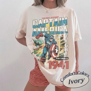 Puede incluir: Una camiseta color marfil con un gr&aacute;fico de Capit&aacute;n Am&eacute;rica en un traje azul, rojo y blanco sosteniendo un escudo. El texto dice THE FIRST AVENGER CAPTAIN AMERICA FIGHT FOR LIBERTY 1941. La camiseta se lleva con pantalones cortos de cuadros rojos y blancos.