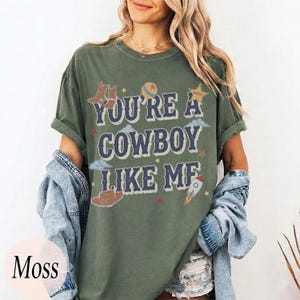 May include: A moss green short-sleeve t-shirt featuring the text "YOU'RE A COWBOY LIKE ME" in dark blue letters. The graphic includes illustrations of cowboy boots, a hat, a ball, a star, clouds, and a rocket ship in various colors.