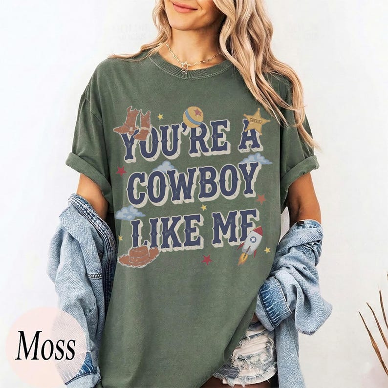 May include: A moss green short-sleeve t-shirt featuring the text "YOU'RE A COWBOY LIKE ME" in dark blue letters. The graphic includes illustrations of cowboy boots, a hat, a ball, a star, clouds, and a rocket ship in various colors.