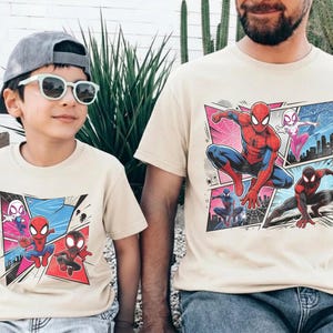K&ouml;nnte beinhalten: Beige T-Shirts mit einem Comic-Grafik-Stil von Spider-Man und anderen Charakteren. Die Shirts werden von einem Kind und einem Erwachsenen getragen. Die Grafik zeigt Spider-Man in verschiedenen Posen mit einem Stadtlandschaftshintergrund.