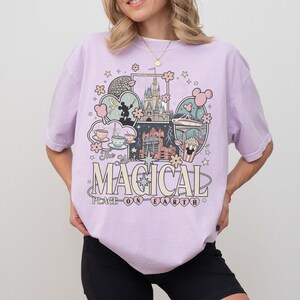 Puede incluir: Una camiseta de color lila claro con un dise&ntilde;o gr&aacute;fico que presenta un castillo, Mickey Mouse y el texto "The Most Magical Place on Earth". El dise&ntilde;o incluye varias ilustraciones y estrellas.