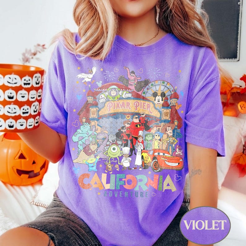 May include: A violet t-shirt featuring a colorful graphic of various Disney Pixar characters and the words "Pixar Pier" and "California Adventure". The shirt has a relaxed fit and short sleeves. A pumpkin-shaped container is held in the left hand.