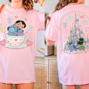 Puede incluir: Camiseta rosa claro con una ilustraci&oacute;n de dibujos animados de Lilo y Stitch. La parte delantera muestra a Stitch y Lilo en una taza de t&eacute; con corazones y globos. La parte trasera muestra una escena de castillo. Dise&ntilde;o colorido.