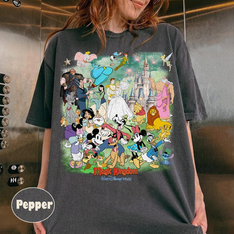 Może przedstawiać: Ciemnoszara koszulka z kolorową grafiką postaci Disneya, w tym Myszki Miki, Kaczora Donalda i Kopciuszka, z napisem "Magic Kingdom" i "Walt Disney World". Projekt zawiera zamek i fajerwerki.