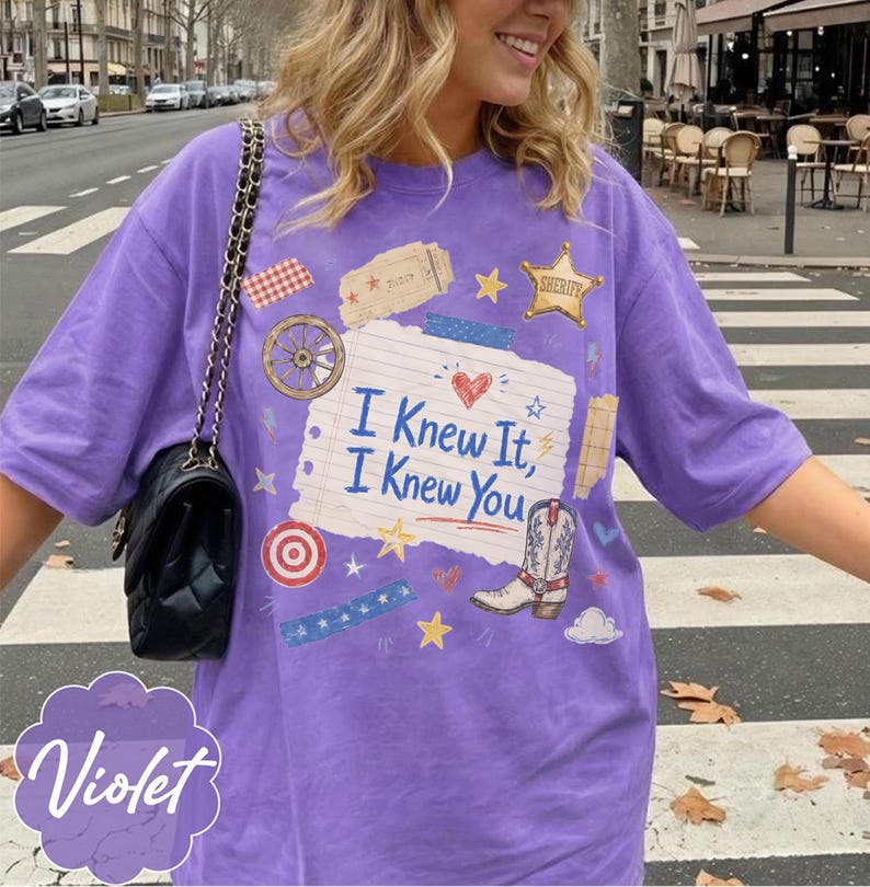 May include: A violet t-shirt featuring a central graphic of a lined notebook paper with the text I Knew It, I Knew You, surrounded by illustrations of a sheriff star, a cowboy boot, a wagon wheel, a target, and stars. A black shoulder bag is worn.