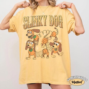 Op de afbeelding: Mosterdgeel T-shirt met de tekst "SLINKY DOG" in een retro lettertype boven een cartoonafbeelding van het Slinky Dog-personage. Het shirt heeft ook de woorden "COMFORT COLOR" en "Mustard" in een cirkelvormig ontwerp.
