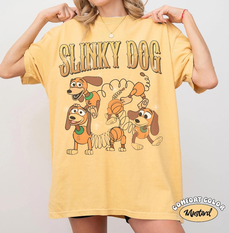Op de afbeelding: Mosterdgeel T-shirt met de tekst "SLINKY DOG" in een retro lettertype boven een cartoonafbeelding van het Slinky Dog-personage. Het shirt heeft ook de woorden "COMFORT COLOR" en "Mustard" in een cirkelvormig ontwerp.