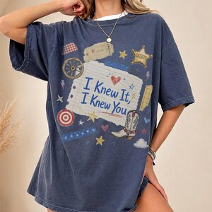 May include: A dark blue short-sleeve t-shirt featuring a central graphic of a lined paper scrap with the text "I Knew It, I Knew You" in blue. The design includes a cowboy boot, a sheriff star, a wagon wheel, and various small stars and hearts.