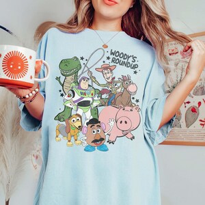 May include: Light blue t-shirt featuring a graphic of characters from the movie Toy Story, including Woody, Buzz Lightyear, and Rex. The text "Woody's Roundup" is printed above the characters. The shirt has short sleeves.
