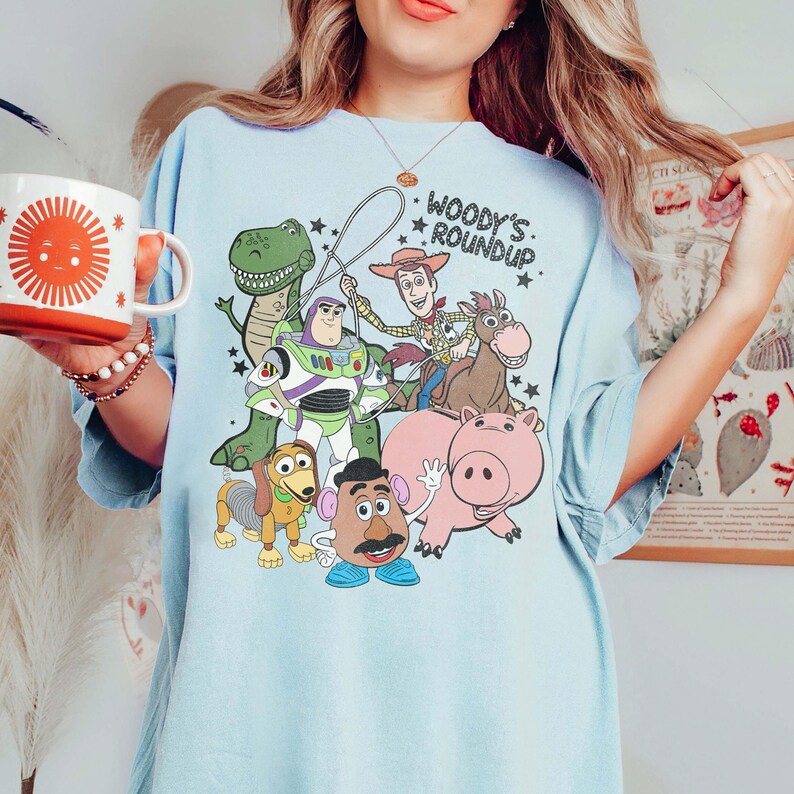 May include: Light blue t-shirt featuring a graphic of characters from the movie Toy Story, including Woody, Buzz Lightyear, and Rex. The text "Woody's Roundup" is printed above the characters. The shirt has short sleeves.