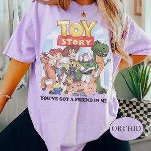 Puede incluir: Camiseta color orqu&iacute;dea con un gr&aacute;fico de personajes de la pel&iacute;cula animada "Toy Story". El dise&ntilde;o incluye a Woody, Buzz Lightyear y otros personajes, con el texto "TOY STORY" y "YOU'VE GOT A FRIEND IN ME". La camiseta tiene mangas cortas.