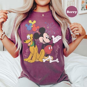 May include: Berry-colored t-shirt featuring a graphic of Mickey Mouse and Pluto with fireworks and balloons. Mickey Mouse is wearing red shorts and Pluto is yellow. The text "Mickey Mouse" is written on the shirt.