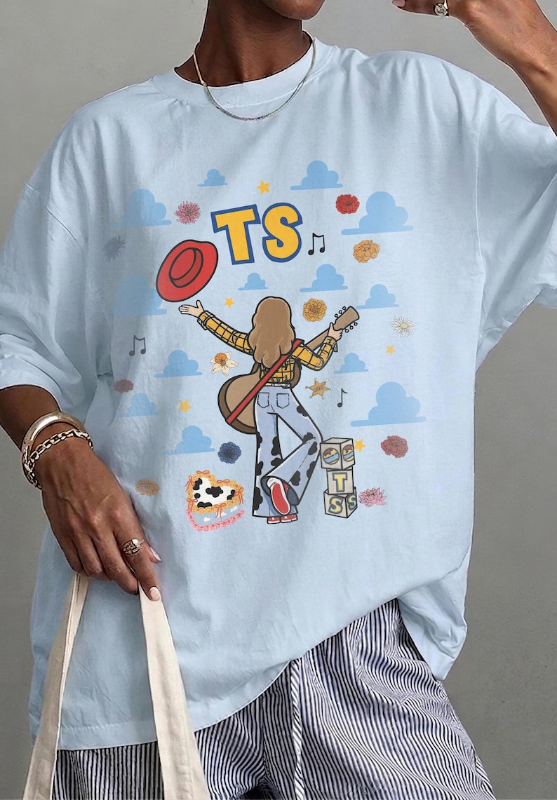 Op de afbeelding: Een lichtblauw t-shirt met een afbeelding van een persoon met lang haar die een gitaar en een rode hoed vasthoudt. Het ontwerp bevat de tekst TS, blauwe wolken, bloemen, muzieknoten en een stapel blokken met de letters T en S.