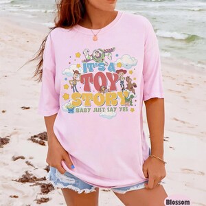 May include: A light pink t-shirt featuring a graphic print of Toy Story characters including Woody, Buzz Lightyear, Jessie, and Bullseye. The text reads IT'S A TOY STORY BABY JUST SAY YES, surrounded by colorful stars and clouds in a retro design.