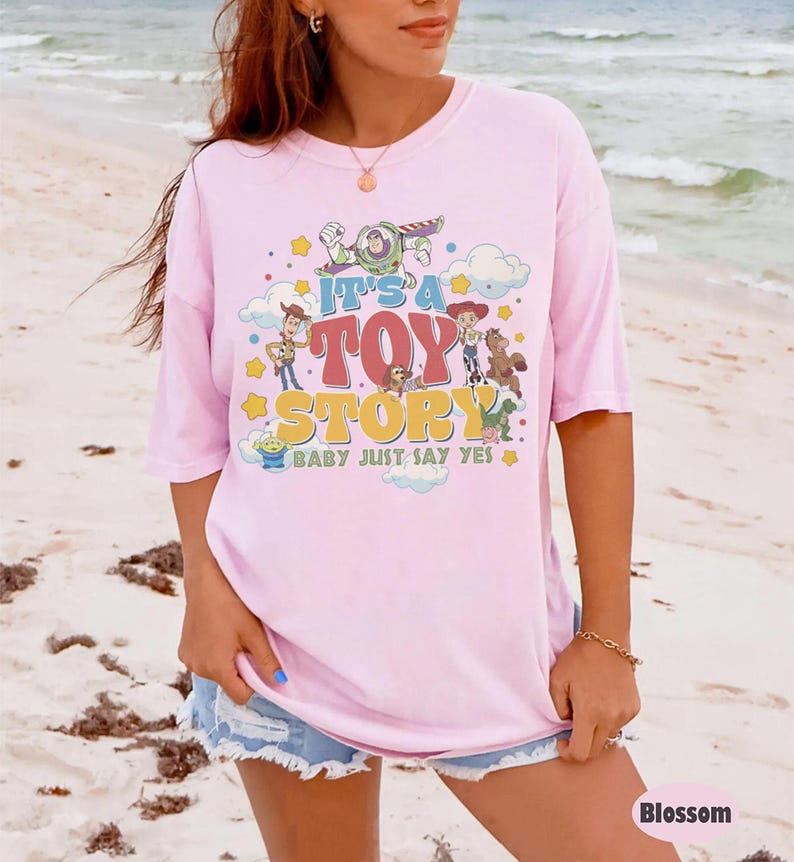 May include: A light pink t-shirt featuring a graphic print of Toy Story characters including Woody, Buzz Lightyear, Jessie, and Bullseye. The text reads IT'S A TOY STORY BABY JUST SAY YES, surrounded by colorful stars and clouds in a retro design.