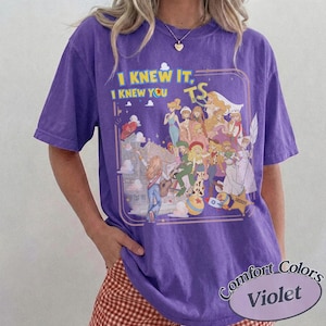 May include: A purple short-sleeve t-shirt featuring a graphic print of various animated characters in a collage style. The text "I KNEW IT, I KNEW YOU TS" is printed in yellow and blue above the characters. The design includes a sheriff star.